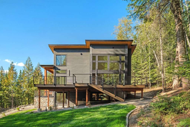 parade-house-42 Modern two-story house with large windows, wood and stone accents, and a sloped roof. It is surrounded by a lush green lawn and tall trees under a clear blue sky. A set of stairs leads up to the main entrance, and a gravel path borders the grass.