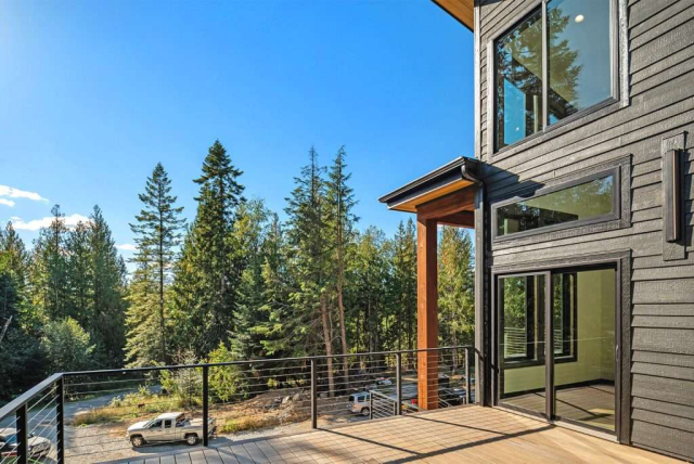 parade-house-44 Modern house exterior with large glass windows and wooden accents on a sunny day. A spacious balcony overlooks a forested area with tall evergreen trees. A road and parked cars are visible in the background.