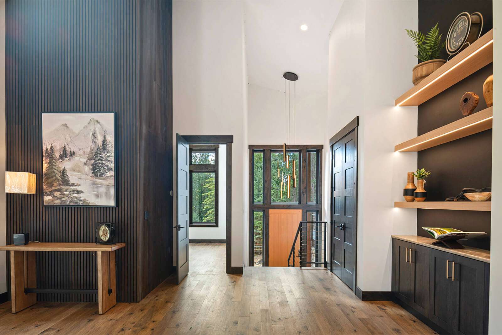 parade-house-6 A modern living space with wooden floors and a high ceiling. There's a landscape painting on a black paneled wall, a wooden console table, and a door. Built-in shelves display decor items, and sunlight streams through a large window.