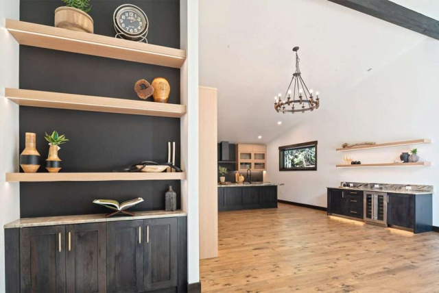 parade-house-8 Modern kitchen with wooden floor, light wood cabinets, and dark lower cabinets. An island features a marble countertop with a potted plant. Built-in shelves display books and decor, and warm lighting creates an inviting atmosphere.